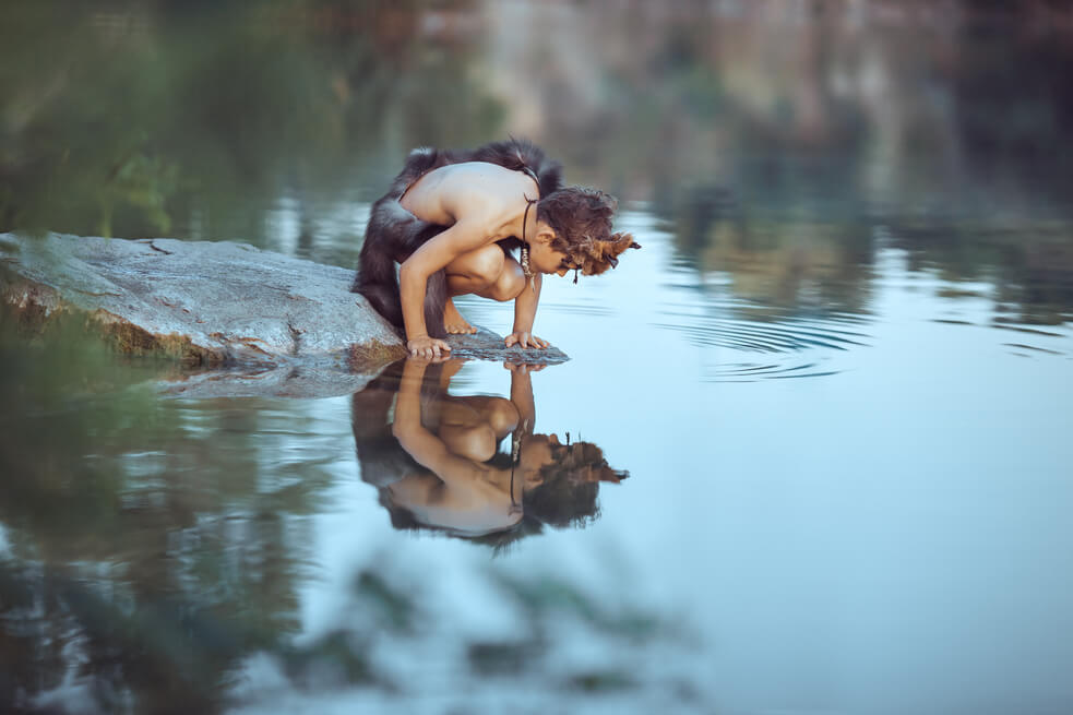 Imagen de niño vestido con pieles mirando su reflejo en un río